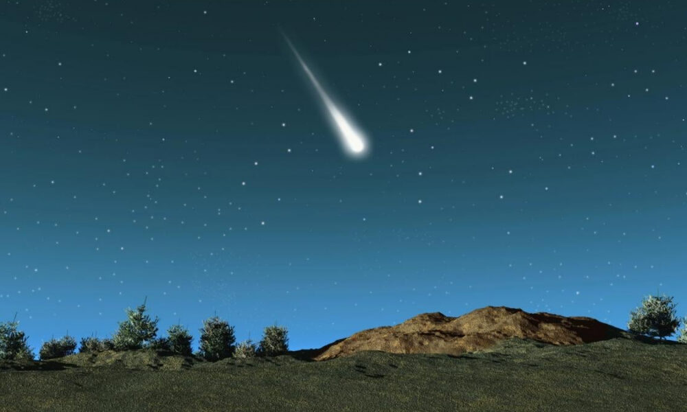 Meteor streaking across a starry night sky above a rocky hill and trees on the horizon.
