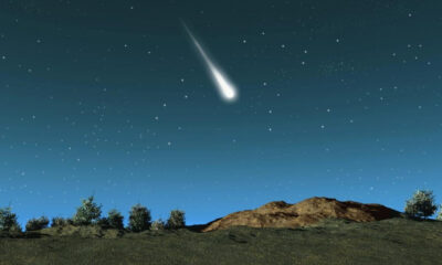 Meteor streaking across a starry night sky above a rocky hill and trees on the horizon.