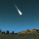 Meteor streaking across a starry night sky above a rocky hill and trees on the horizon.