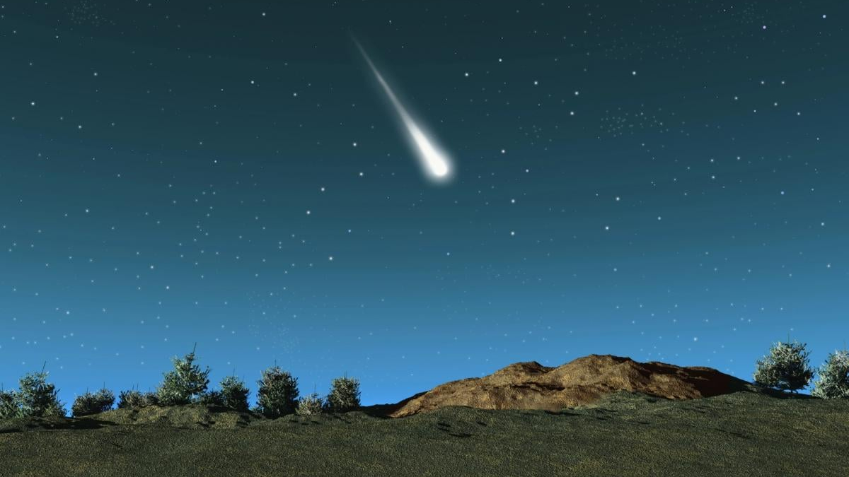 Meteor streaking across a starry night sky above a rocky hill and trees on the horizon.