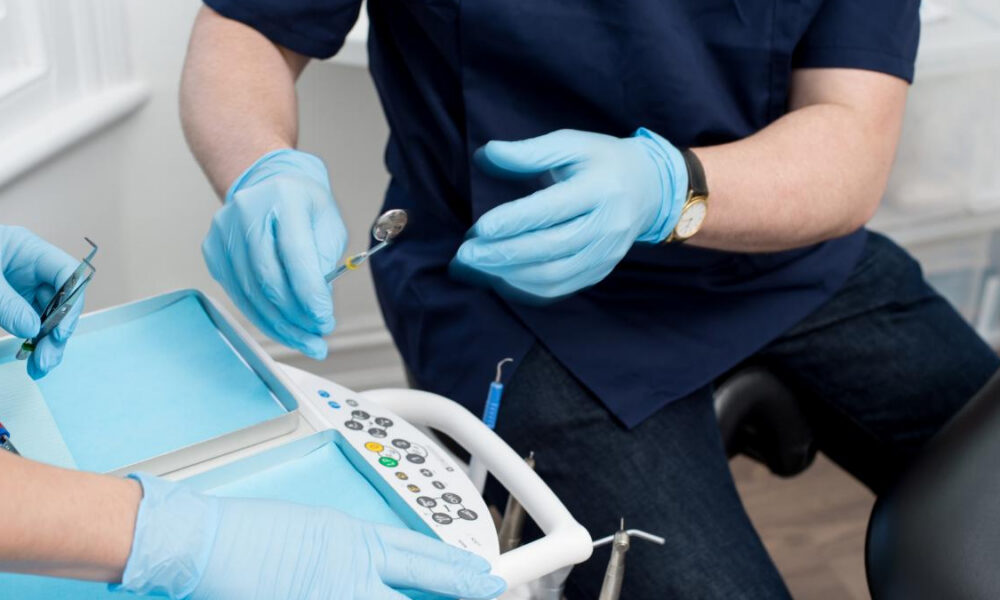 Dentist in blue scrubs and gloves handling dental drill over a patient chair with tray and controls nearby.