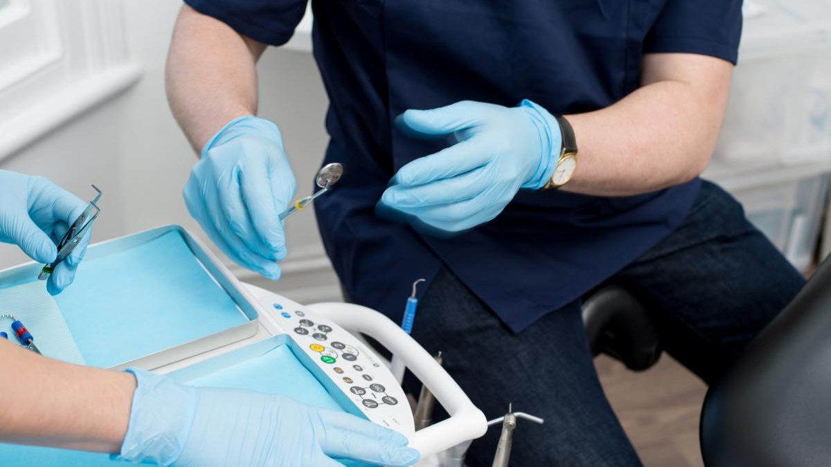 Dentist in blue scrubs and gloves handling dental drill over a patient chair with tray and controls nearby.
