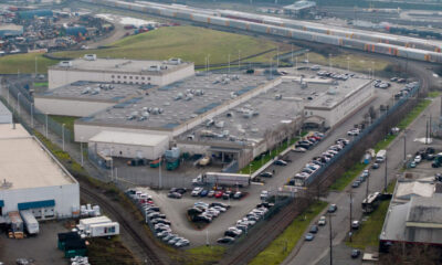 Aerial view of a large industrial complex with flat rooftops, parking lots, and nearby rail tracks and freight trains.