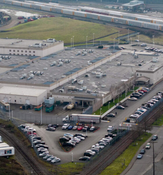 Aerial view of a large industrial complex with flat rooftops, parking lots, and nearby rail tracks and freight trains.