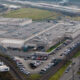 Aerial view of a large industrial complex with flat rooftops, parking lots, and nearby rail tracks and freight trains.