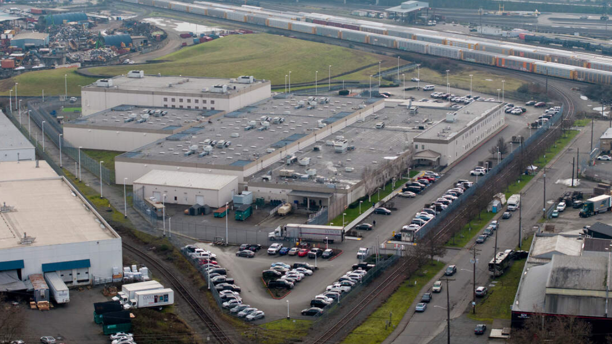 Aerial view of a large industrial complex with flat rooftops, parking lots, and nearby rail tracks and freight trains.