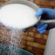 Gloved hand pours granulated sugar from a metal pan into a pot, sugar falling midair into a kitchen pot.