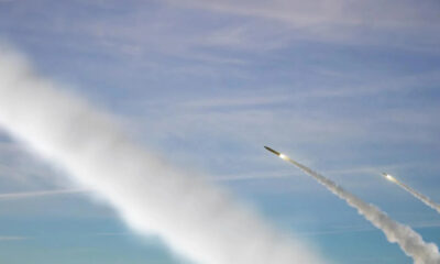 Two rockets angled upward, leaving white exhaust trails across a blue sky.
