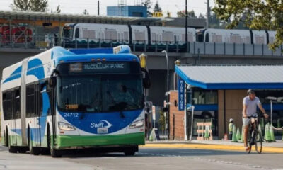 Blue-and-white Swift transit bus driving on a street as a cyclist passes by; an elevated train line and station are in the background.