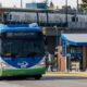 Blue-and-white Swift transit bus driving on a street as a cyclist passes by; an elevated train line and station are in the background.