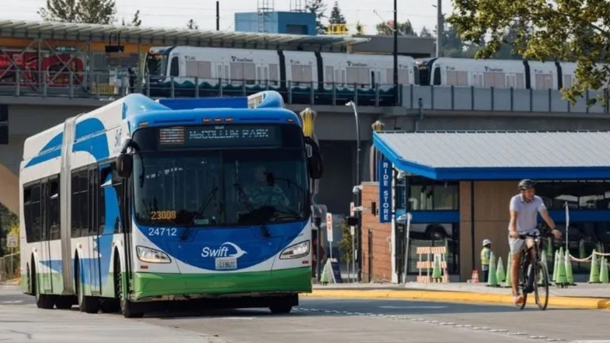 Blue-and-white Swift transit bus driving on a street as a cyclist passes by; an elevated train line and station are in the background.