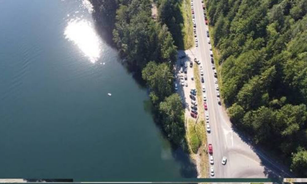 Aerial view of a blue lake with a tree-lined shore beside a busy road and line of parked cars on the edge of a forested area.