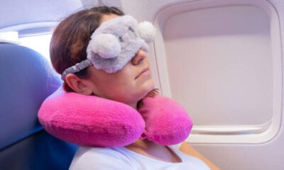 Traveler resting on a plane seat with a soft gray eye mask and two pink inflatable neck pillows, by the window.