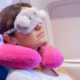 Traveler resting on a plane seat with a soft gray eye mask and two pink inflatable neck pillows, by the window.