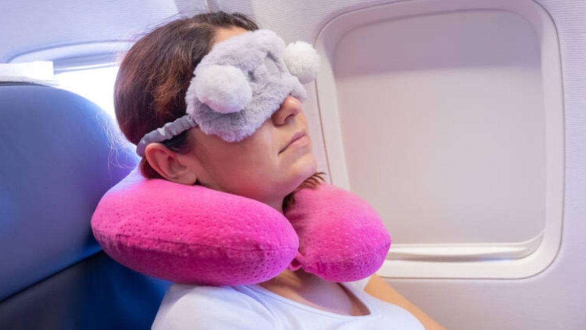 Traveler resting on a plane seat with a soft gray eye mask and two pink inflatable neck pillows, by the window.