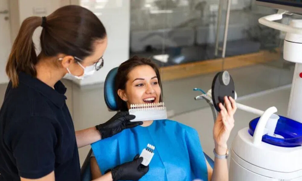 Dental assistant in gloves and mask shows a tooth color shade guide to a smiling patient in a dental chair.