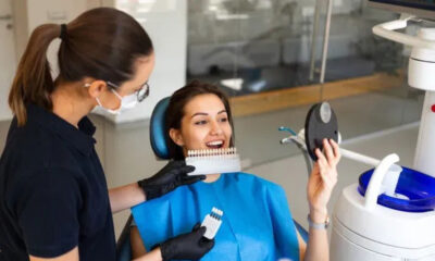 Dental assistant in gloves and mask shows a tooth color shade guide to a smiling patient in a dental chair.