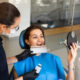Dental assistant in gloves and mask shows a tooth color shade guide to a smiling patient in a dental chair.