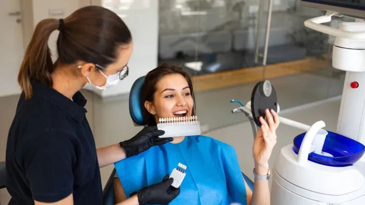 Dental assistant in gloves and mask shows a tooth color shade guide to a smiling patient in a dental chair.