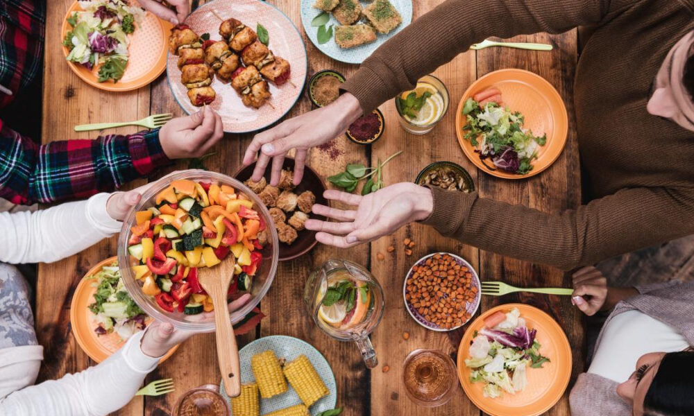 Overhead view of a group sharing a colorful salad and various dishes on a wooden table, hands reaching for food.