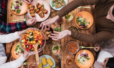 Overhead view of a group sharing a colorful salad and various dishes on a wooden table, hands reaching for food.