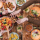 Overhead view of a group sharing a colorful salad and various dishes on a wooden table, hands reaching for food.