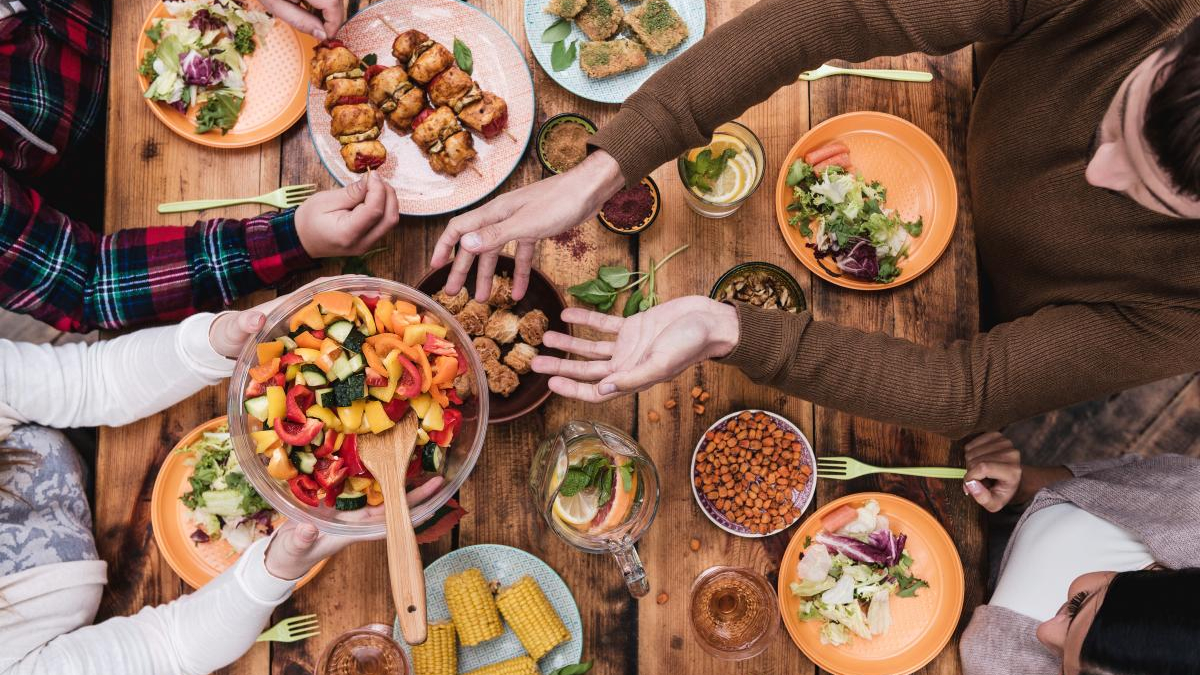 Overhead view of a group sharing a colorful salad and various dishes on a wooden table, hands reaching for food.
