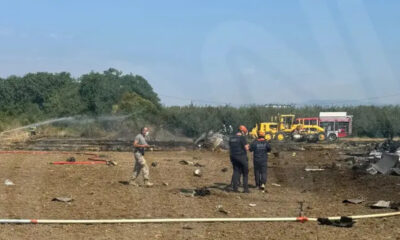 Emergency responders spray water on a burned field with scattered debris and emergency vehicles in the background, daytime scene.