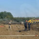 Emergency responders spray water on a burned field with scattered debris and emergency vehicles in the background, daytime scene.