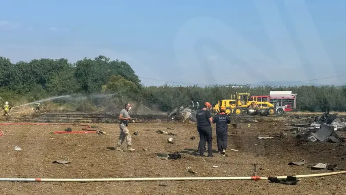 Emergency responders spray water on a burned field with scattered debris and emergency vehicles in the background, daytime scene.