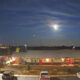 Nighttime view of a single-story building with a parking lot and cars, under a cloudy sky with a bright moon.