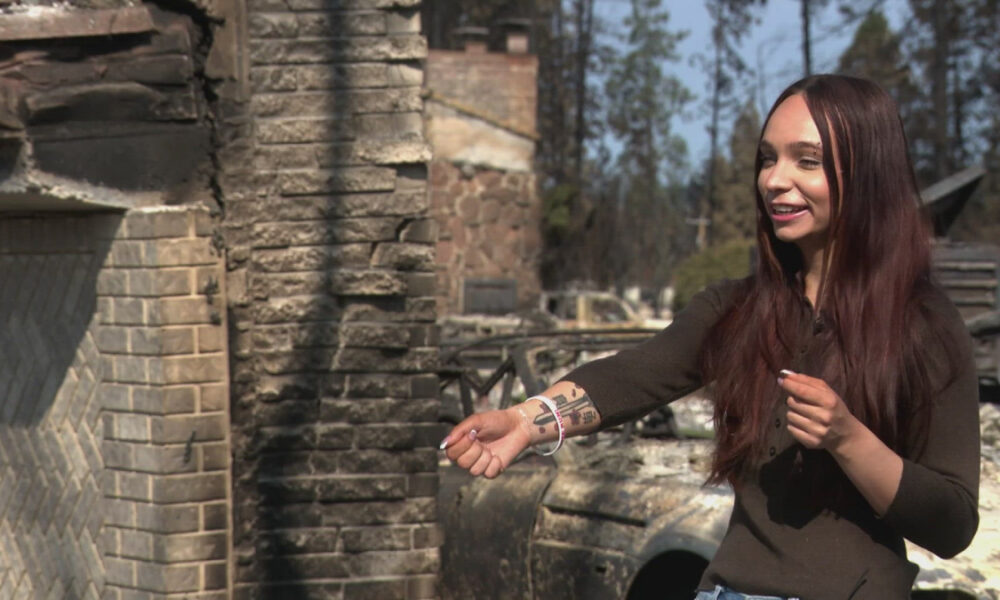 Woman with long dark hair and tattooed forearm extends her arm, holding a cigarette, in front of charred rubble from a fire.