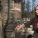 Woman with long dark hair and tattooed forearm extends her arm, holding a cigarette, in front of charred rubble from a fire.