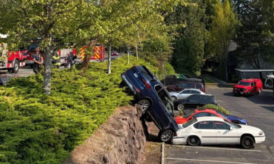 Blue SUV teeters on a rocky ledge above a parking lot, leaning onto a white sedan as responders work nearby.