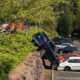 Blue SUV teeters on a rocky ledge above a parking lot, leaning onto a white sedan as responders work nearby.