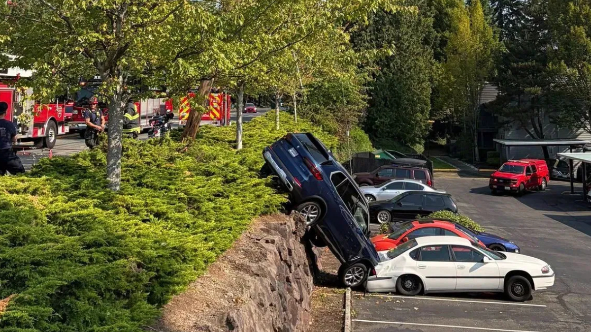 Blue SUV teeters on a rocky ledge above a parking lot, leaning onto a white sedan as responders work nearby.