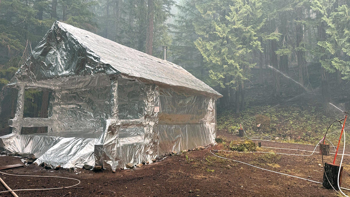Aluminum-foil wrapped shelter in a forest clearing, with hoses running around and a sprinkler spraying mist nearby.