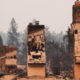 Charred chimneys and burned-out cars in a wildfire-damaged neighborhood, smoke lingering in the air.