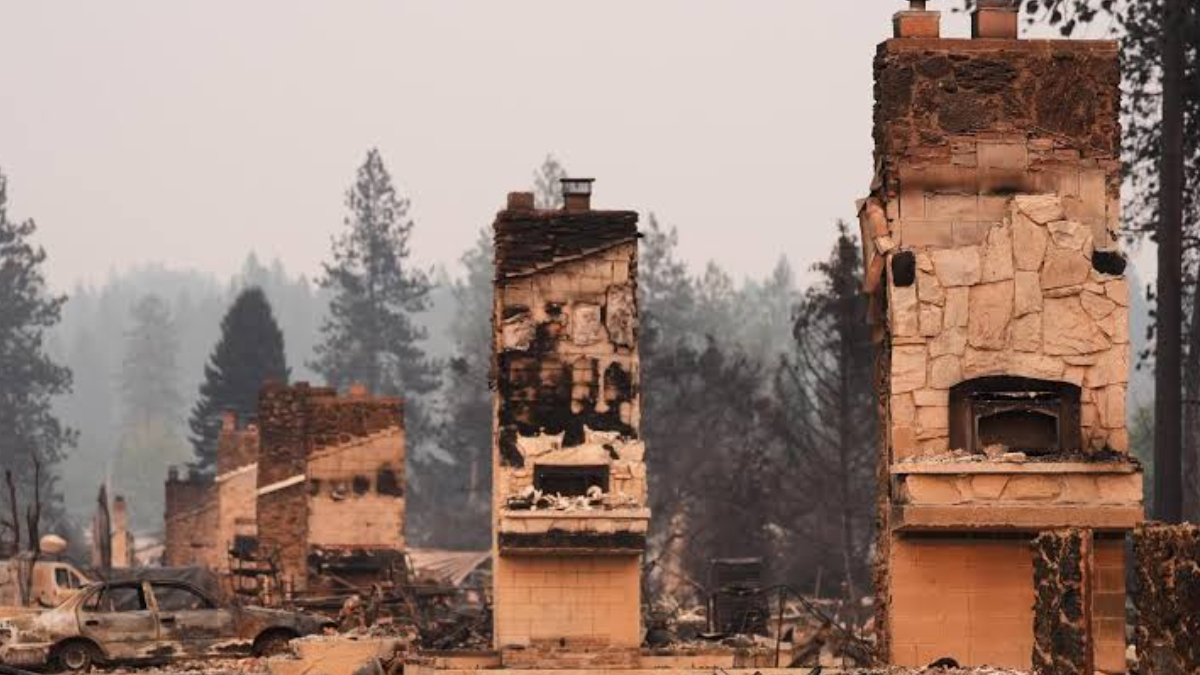 Charred chimneys and burned-out cars in a wildfire-damaged neighborhood, smoke lingering in the air.