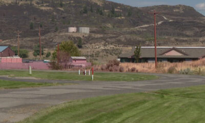 Rural scene featuring a pink wall and buildings, utility poles, and a grassy foreground with hills in the background beyond the road.