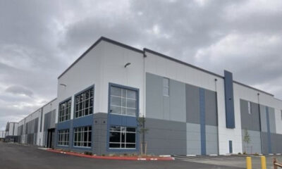 Exterior of a modern industrial warehouse with gray-blue panels and large windows under a cloudy sky, foreground shows a parking area and red curb.