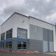 Exterior of a modern industrial warehouse with gray-blue panels and large windows under a cloudy sky, foreground shows a parking area and red curb.