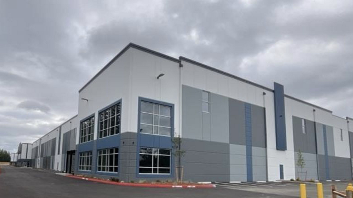 Exterior of a modern industrial warehouse with gray-blue panels and large windows under a cloudy sky, foreground shows a parking area and red curb.