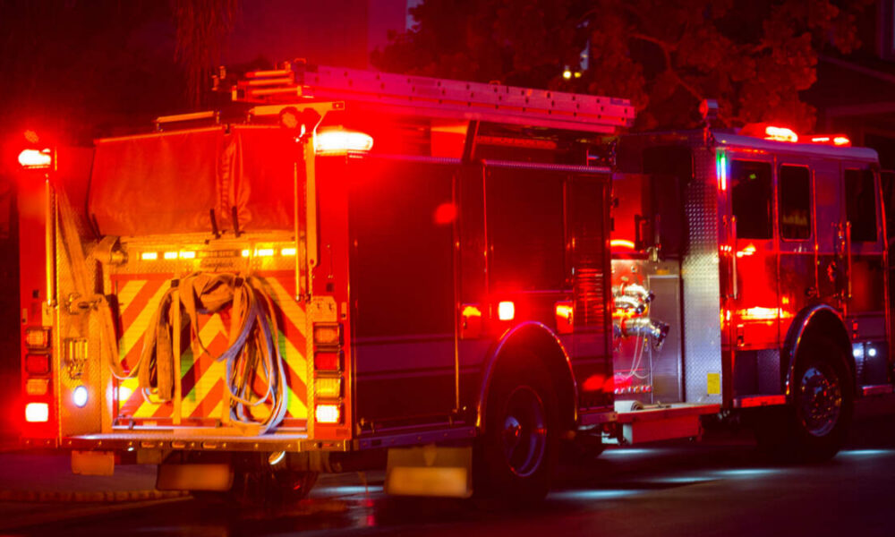 Fire engine parked at night with red emergency lights flashing, hoses coiled at the back and equipment compartments visible on the sides.