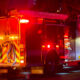 Fire engine parked at night with red emergency lights flashing, hoses coiled at the back and equipment compartments visible on the sides.