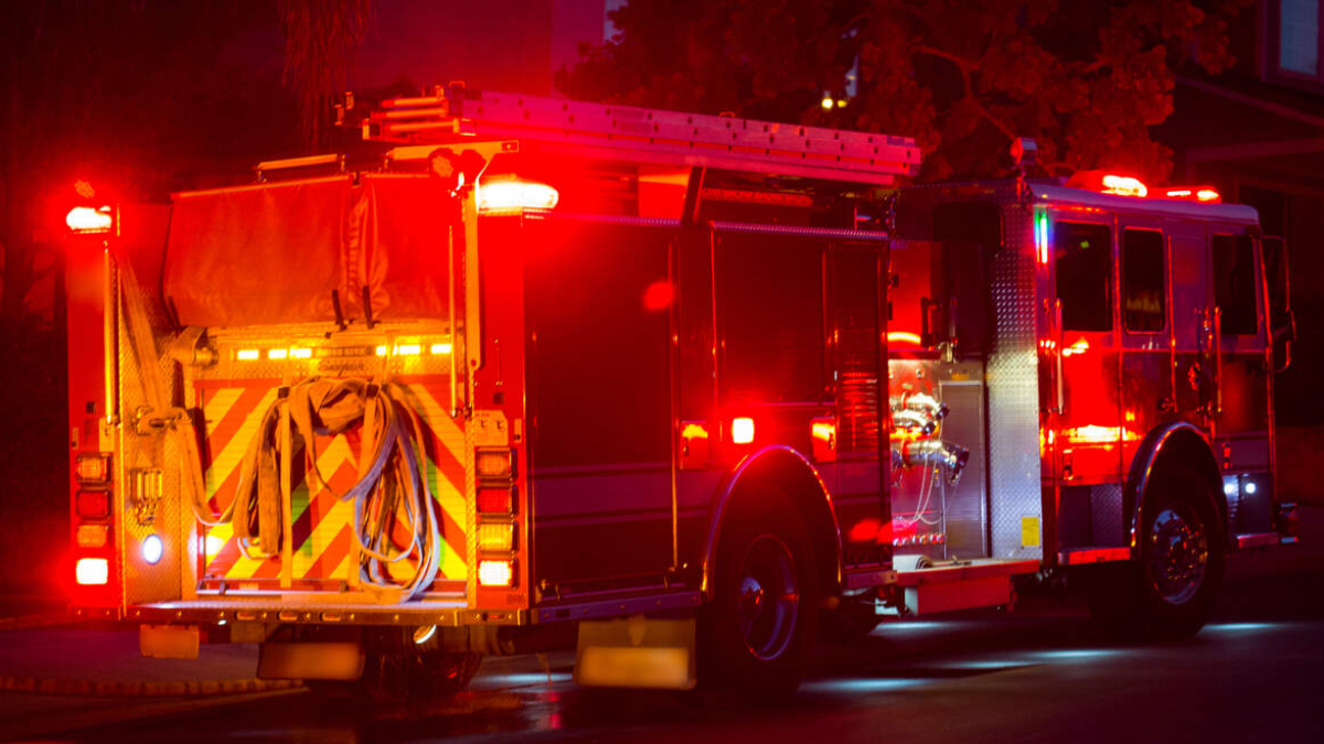 Fire engine parked at night with red emergency lights flashing, hoses coiled at the back and equipment compartments visible on the sides.