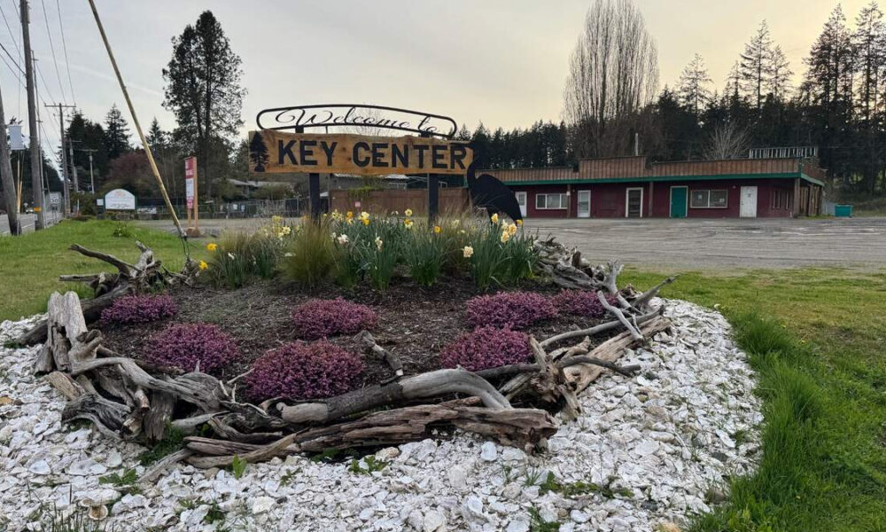 Roundabout flower bed with purple shrubs, yellow daffodils, and driftwood border, featuring a large 'Welcome to Key Center' sign in a small town.