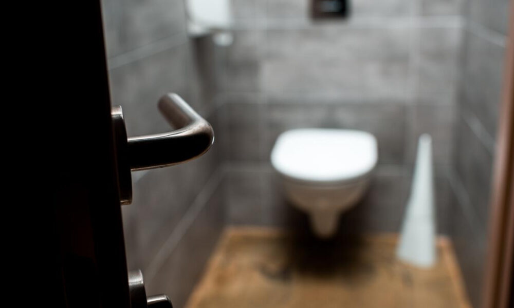 Blurry bathroom with a wall-mounted toilet seen from the doorway; metal door lever in the foreground.