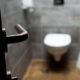 Blurry bathroom with a wall-mounted toilet seen from the doorway; metal door lever in the foreground.