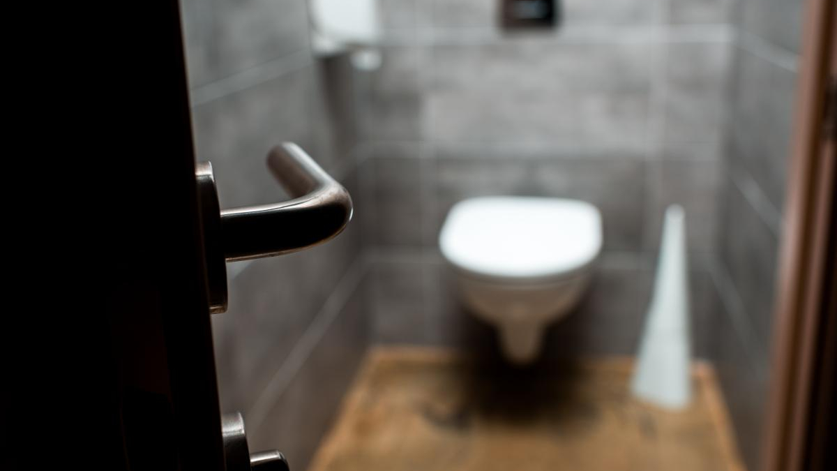Blurry bathroom with a wall-mounted toilet seen from the doorway; metal door lever in the foreground.